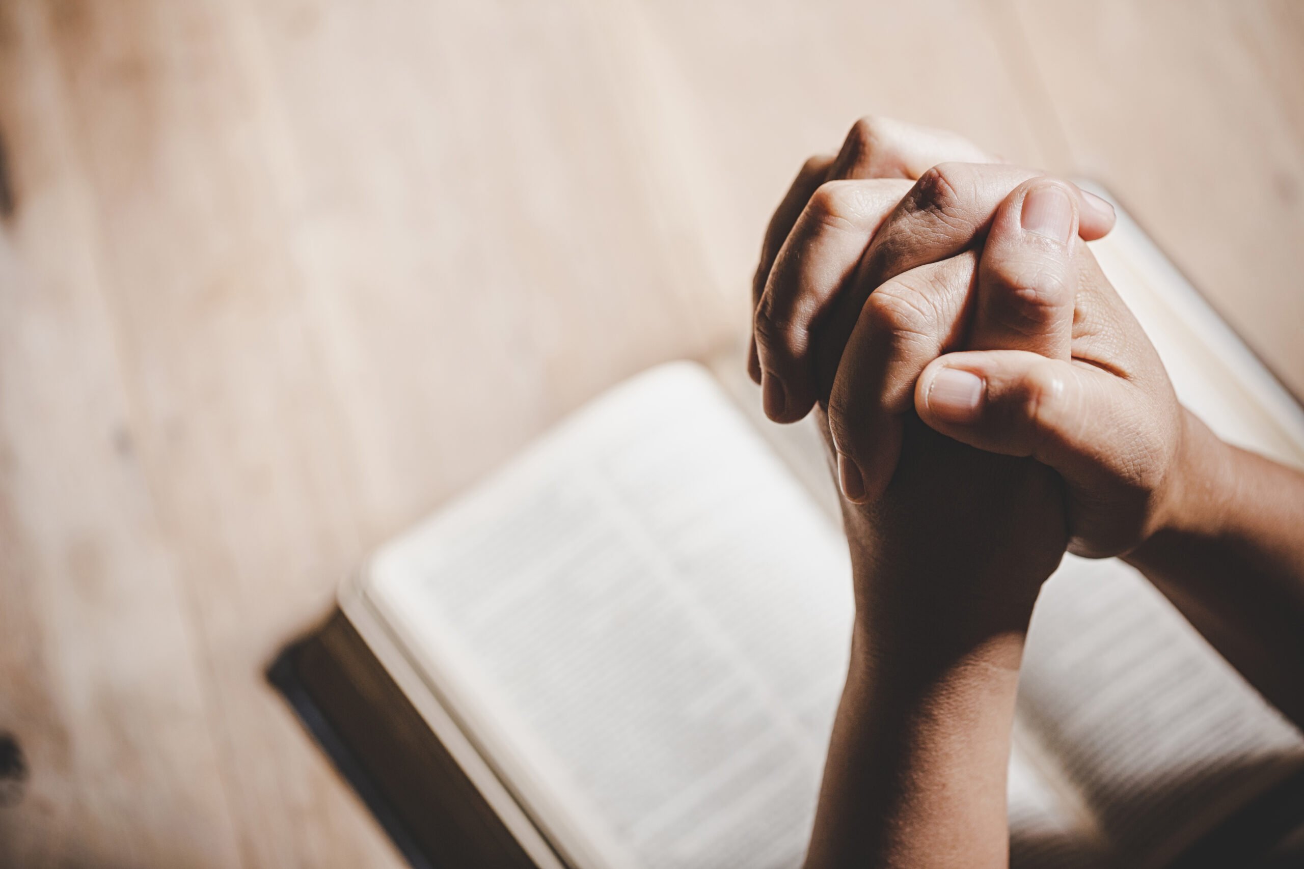 Close-up of hands clasped in prayer over an open Bible on a wooden table.