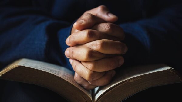 Hands clasped in prayer over an open Bible, with soft lighting and a dark background creating a quiet, reflective mood.