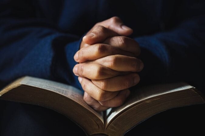 Hands clasped in prayer over an open Bible, with soft lighting and a dark background creating a quiet, reflective mood.
