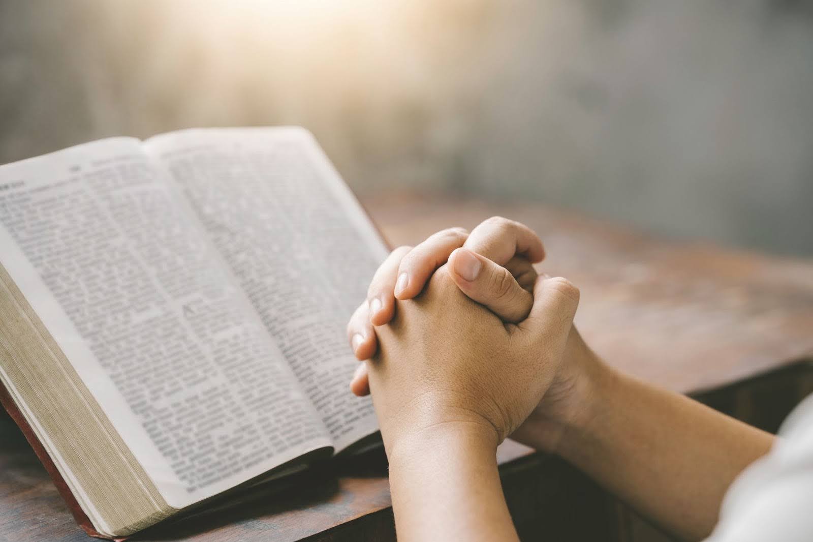 Hands clasped in prayer beside an open Bible on a wooden table in soft morning light.