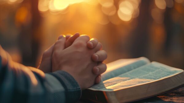 Hands clasped in prayer over an open Bible during a peaceful morning sunrise.