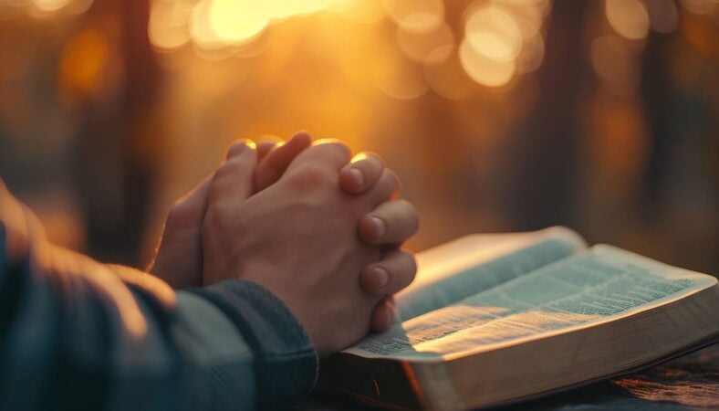 Hands clasped in prayer over an open Bible during a peaceful morning sunrise.