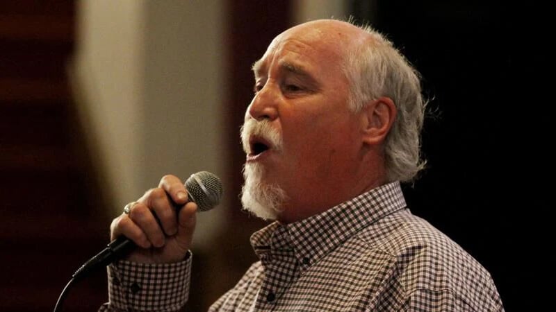 Older man with white hair and a goatee singing into a handheld microphone on stage.