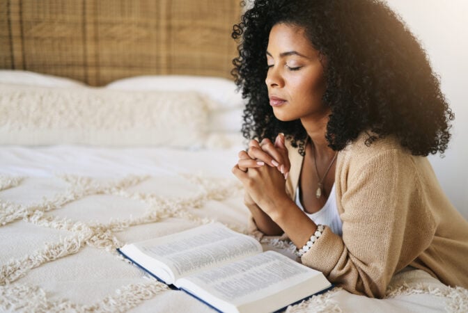 Woman praying quietly on her bed with an open Bible in front of her