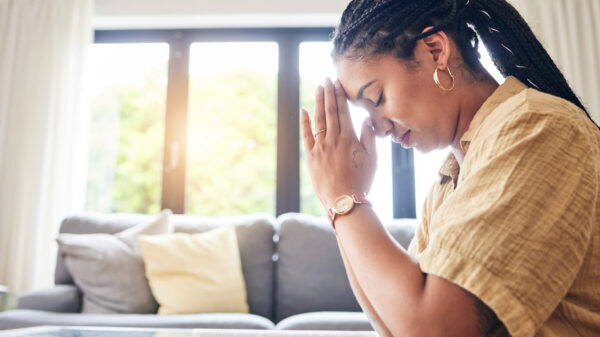 Woman sitting indoors with her head bowed and hands together in prayer, with soft morning light behind her